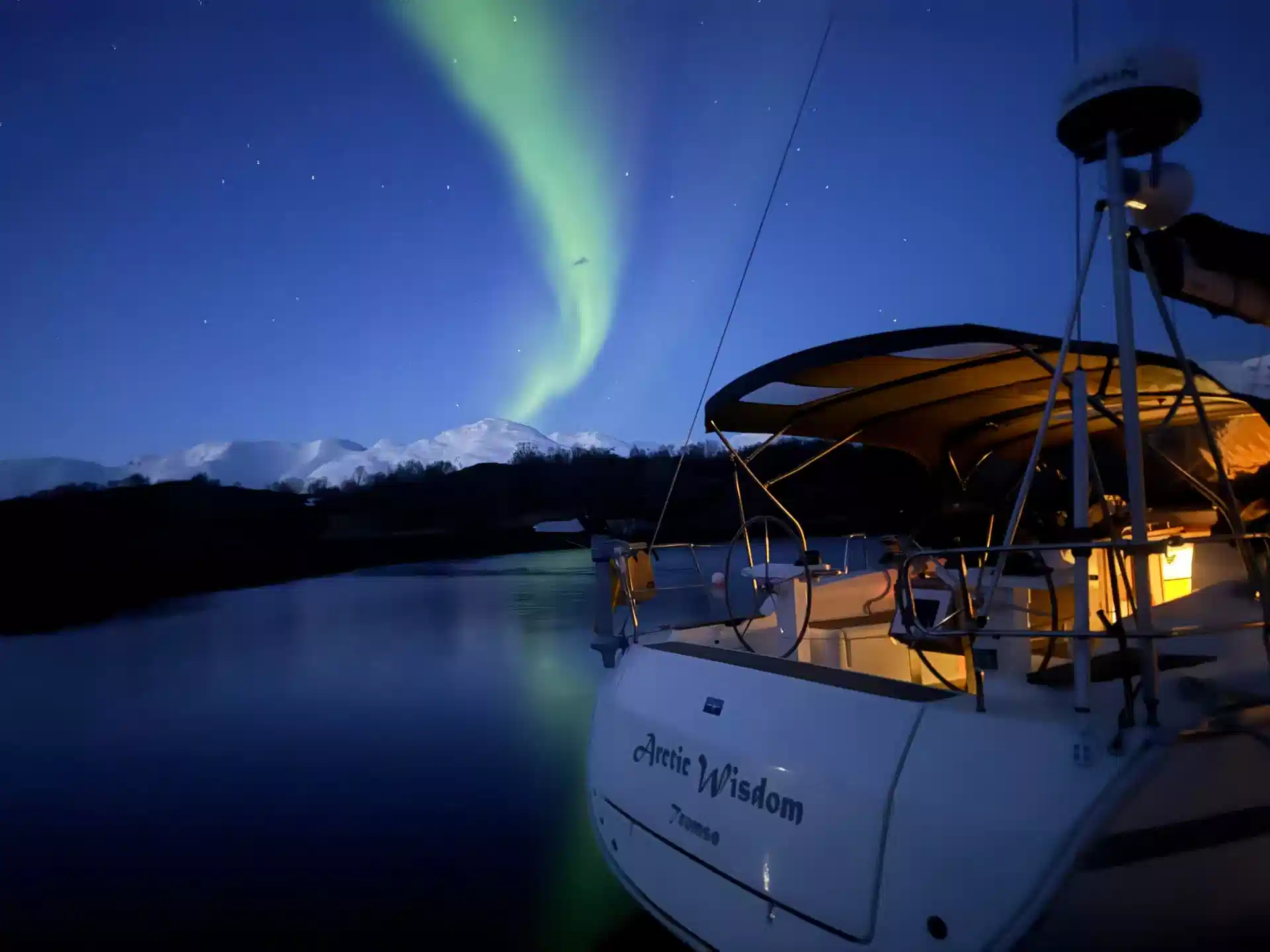Arctic Wisdom lying at anchor in a Norwegian fjord at night, cockpit lit warm beneath the northern lights