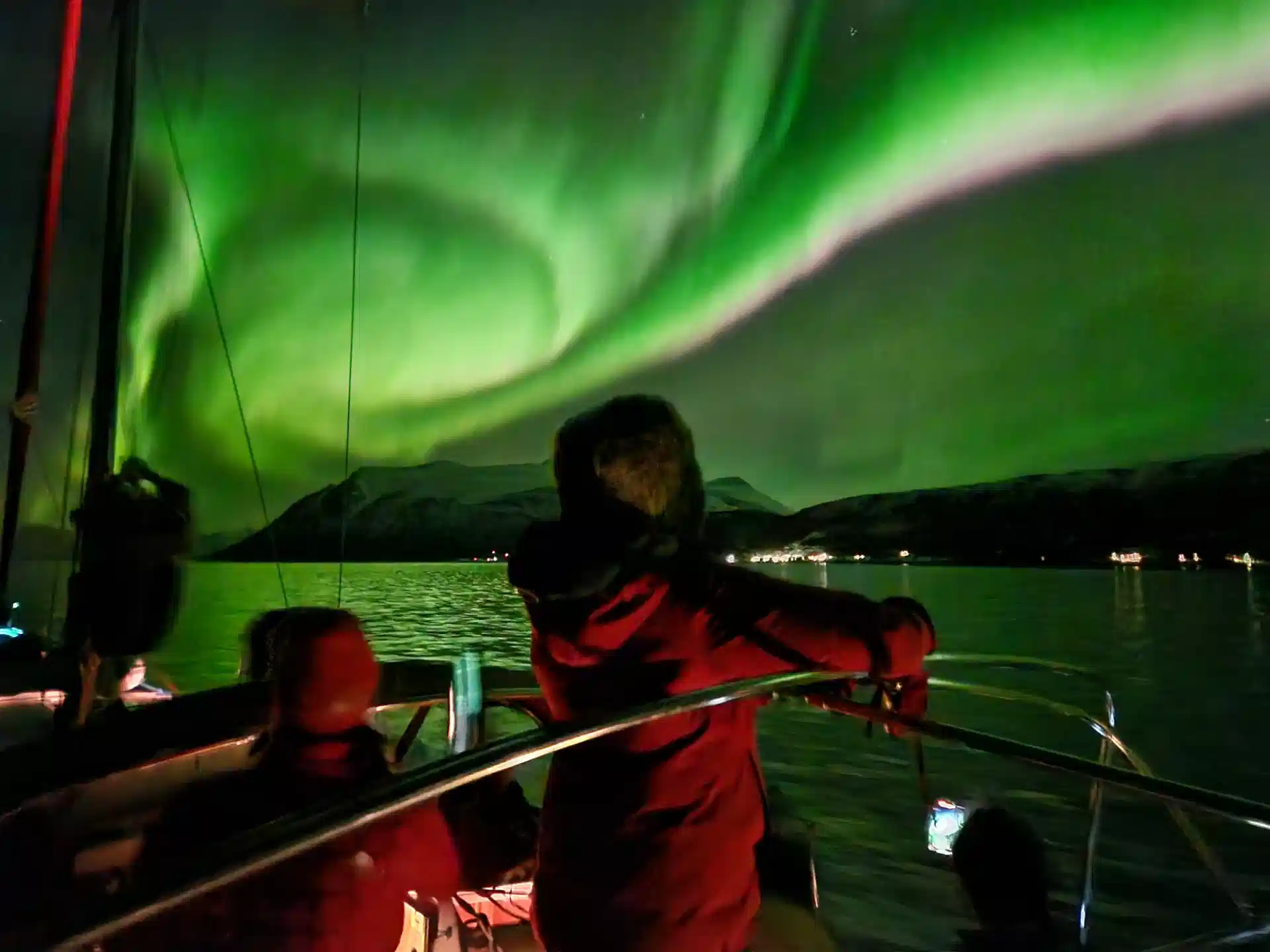 Guests in sailing suits on the deck of a yacht, watching the northern lights over a fjord near Tromsø